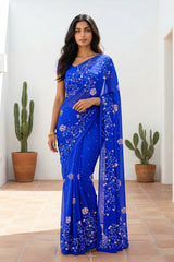 An Indian woman standing in a minimal white courtyard wearing a royal blue chiffon saree embellished with vgolden sliver and pink sequin vines of Stylized Jasmine by Umaid Couture.  
