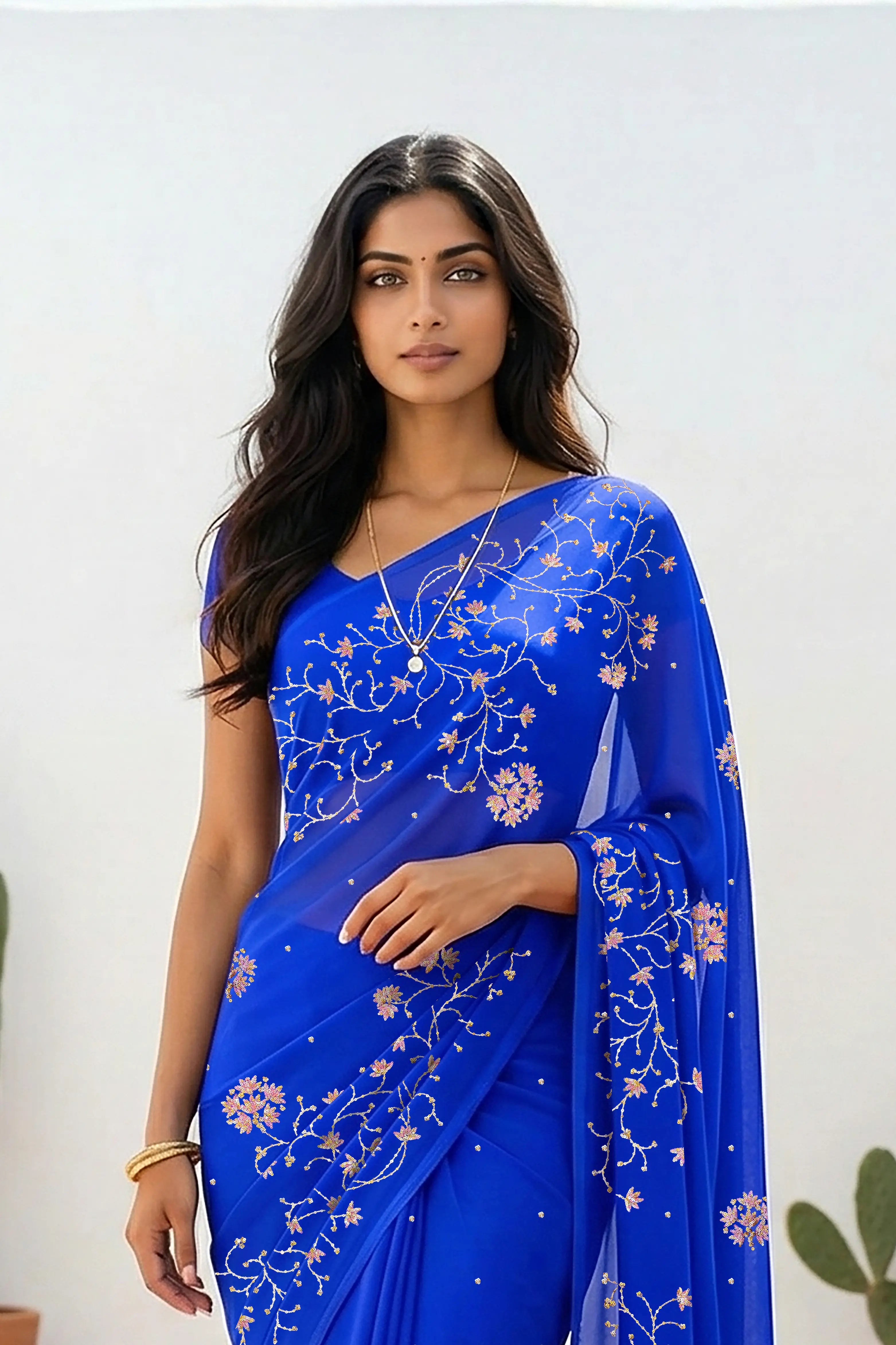 An Indian woman standing in a minimal white courtyard wearing a royal blue chiffon saree embellished with vgolden sliver and pink sequin vines of Stylized Jasmine by Umaid Couture.  
