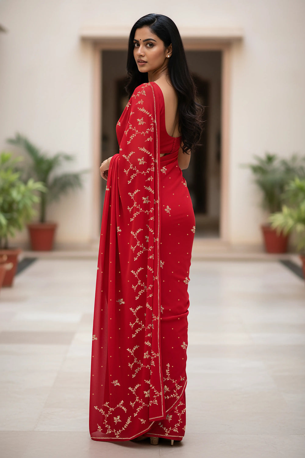An Indian woman standing in a minimal courtyard wearing red color chiffon saree embellished with golden flower pattern vines done using the exquisite Aari-Taari craft  by Umaid Couture.  
