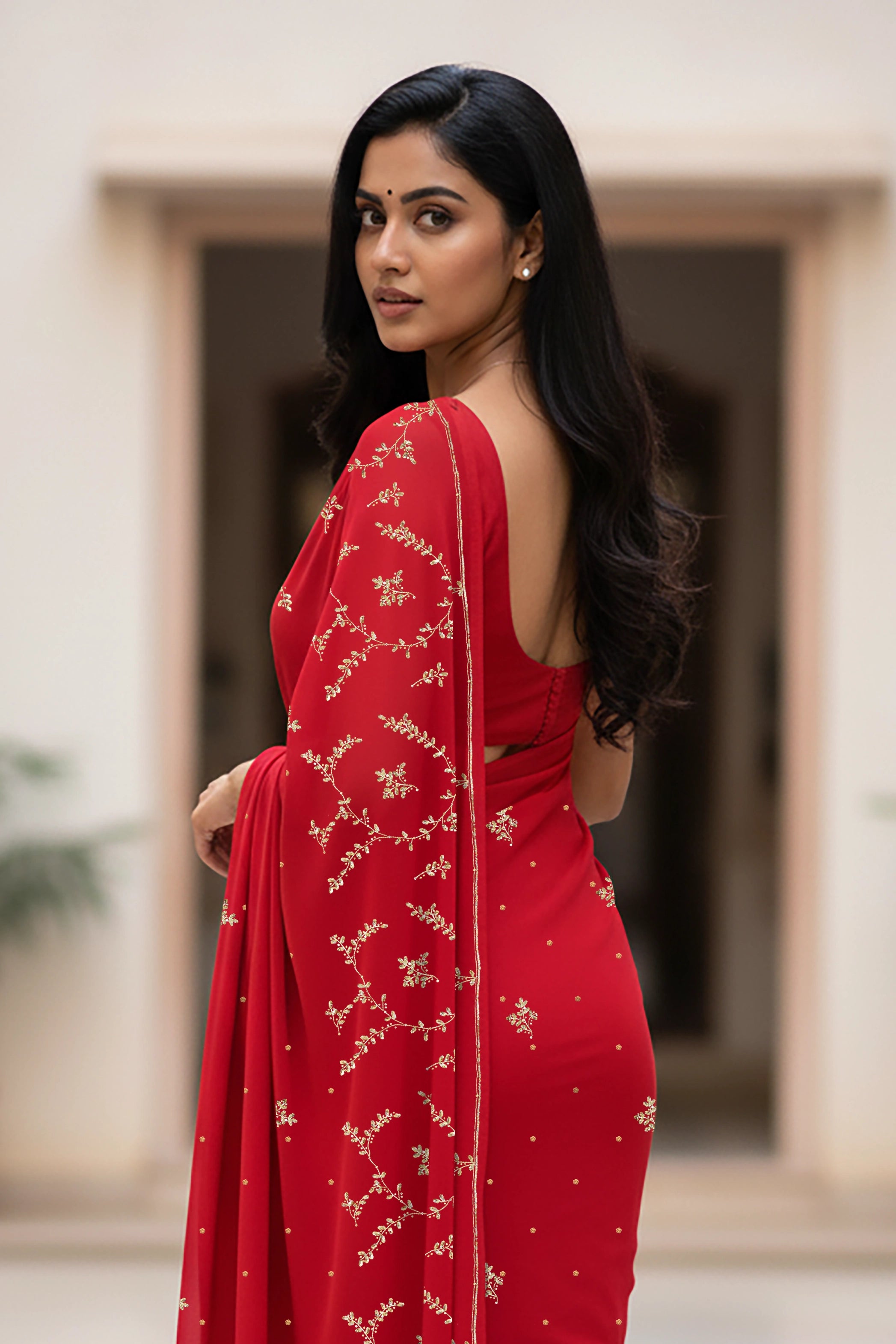 An Indian woman standing in a minimal courtyard wearing red color chiffon saree embellished with golden flower pattern vines done using the exquisite Aari-Taari craft  by Umaid Couture.  
