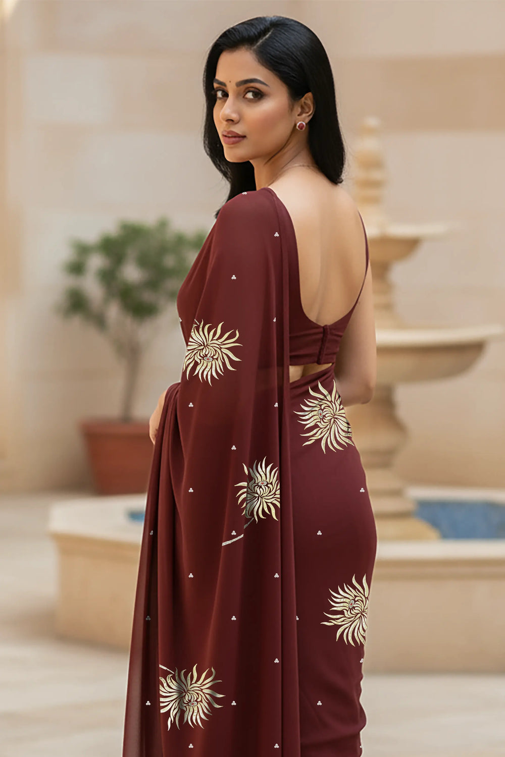 An Indian woman standing in a hotel courtyard wearing a coco brown chiffon saree embellished with shevanti flower motifs made with shimmering Zari, Sequins, and Aari-Zardosi craft by Umaid Couture.  
